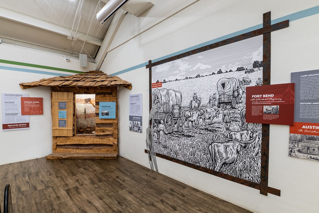 Museum exhibit featuring a wooden frontier cabin facade and a large black-and-white mural of covered wagons crossing a field, with informational panels about Texas history on the surrounding walls.