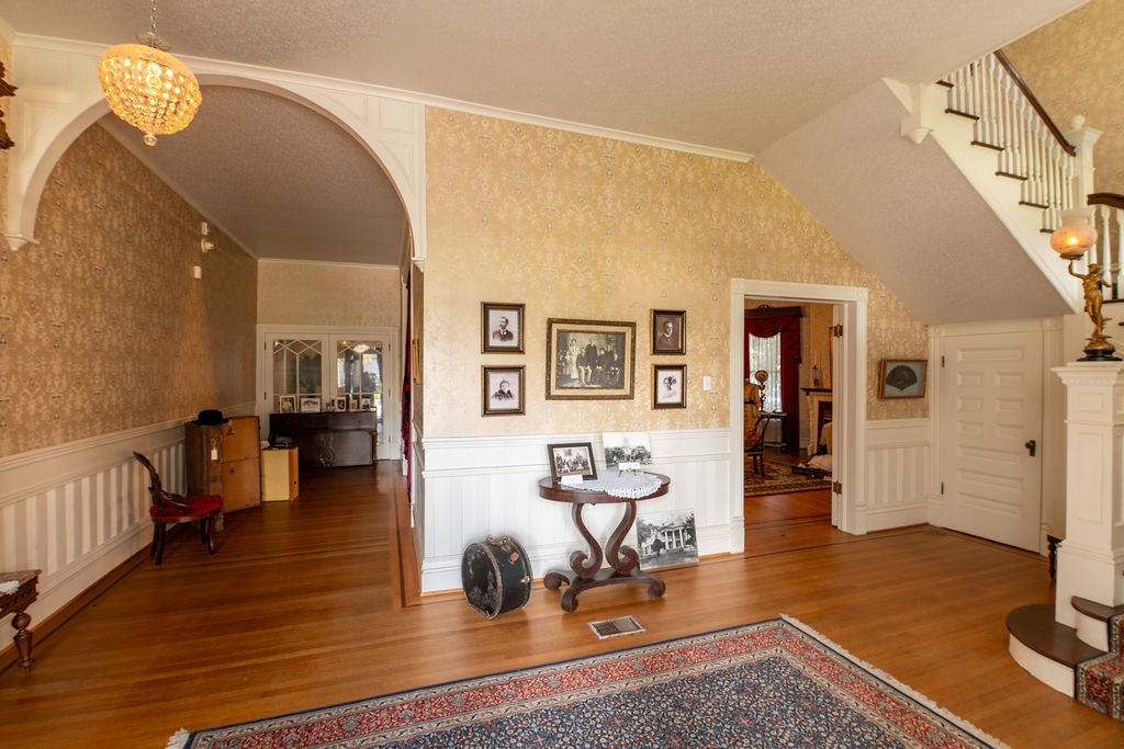 A spacious, vintage-style foyer with wood floors, ornate wallpaper, family photos on the wall, a round table with framed pictures, a staircase, archway, chandelier, and a view into adjacent rooms.