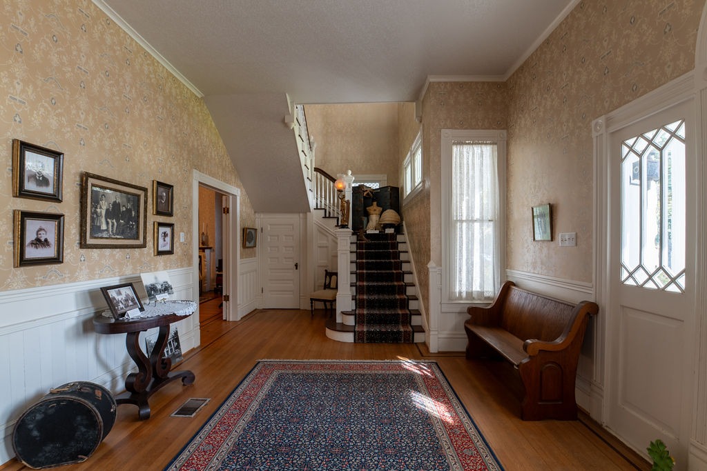 A vintage foyer with wood floors, a patterned rug, a wooden bench, table, framed photos on wallpapered walls, and a carpeted staircase leading upstairs. Sunlight streams through a door with decorative glass.