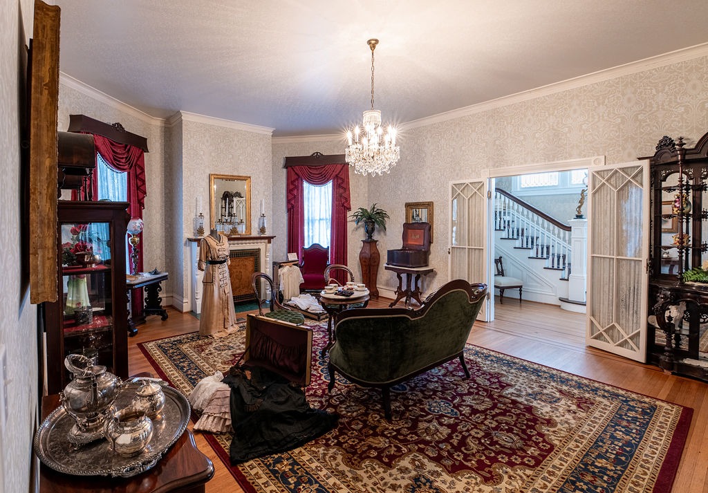 A vintage parlor with ornate furniture, a chandelier, a fireplace, elegant drapes, and a decorative rug. Mannequins in period clothing and antique decor add a historical touch. A staircase is visible through open doors.