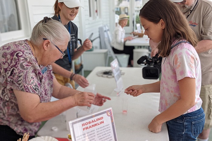 An older woman helps a young girl with a science activity at a table labeled "Rosalind Franklin." Other people and activity materials are visible in the background on a covered porch.