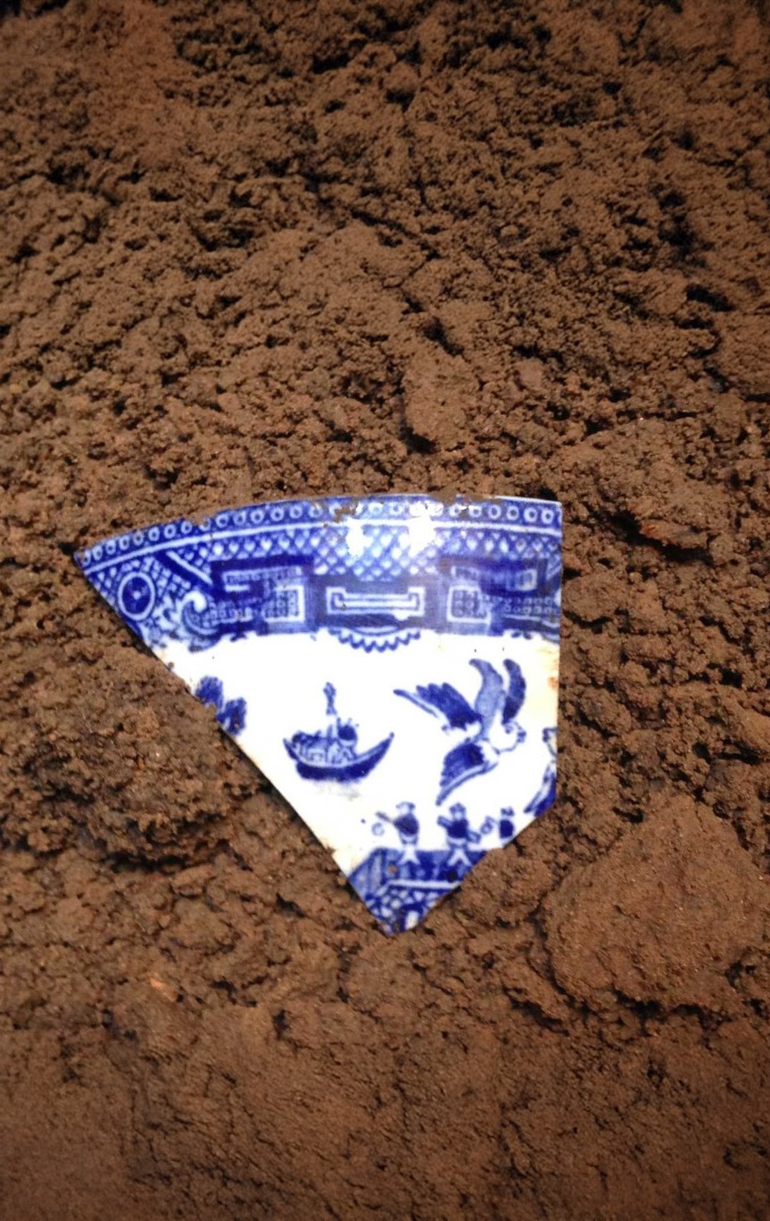 A blue and white porcelain shard with a painted scene, partially buried in brown soil. The shard features birds, water, and architectural designs in traditional style.