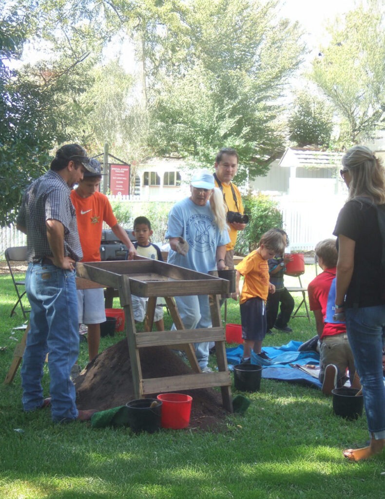 A group of children and adults participate in an outdoor hands-on activity, possibly sifting dirt or sand through a wooden frame. They are on green grass with trees and buildings in the background.