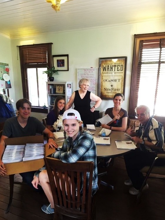 A group of six people sit and stand around a table in a cozy room, working with papers and envelopes. A box of mail is on the table, and a "WANTED" poster is on the wall behind them. They appear cheerful and collaborative.
