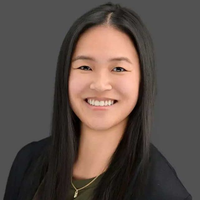 A woman with long straight black hair, wearing a dark top and a gold necklace, smiles at the camera against a plain gray background.