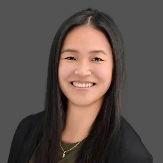 A woman with long straight black hair, wearing a dark top and a gold necklace, smiles at the camera against a plain gray background.