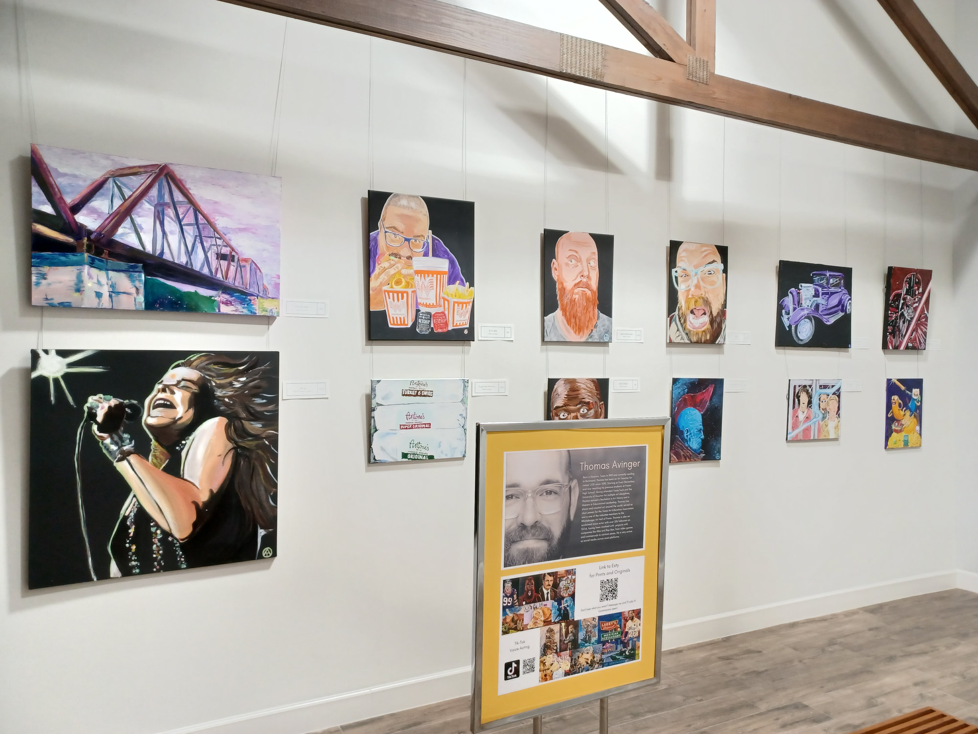 A gallery wall displays colorful paintings, including portraits, a singer, a bridge, cars, and pop culture art. Wooden beams and a prominent artist information board are visible in the foreground.