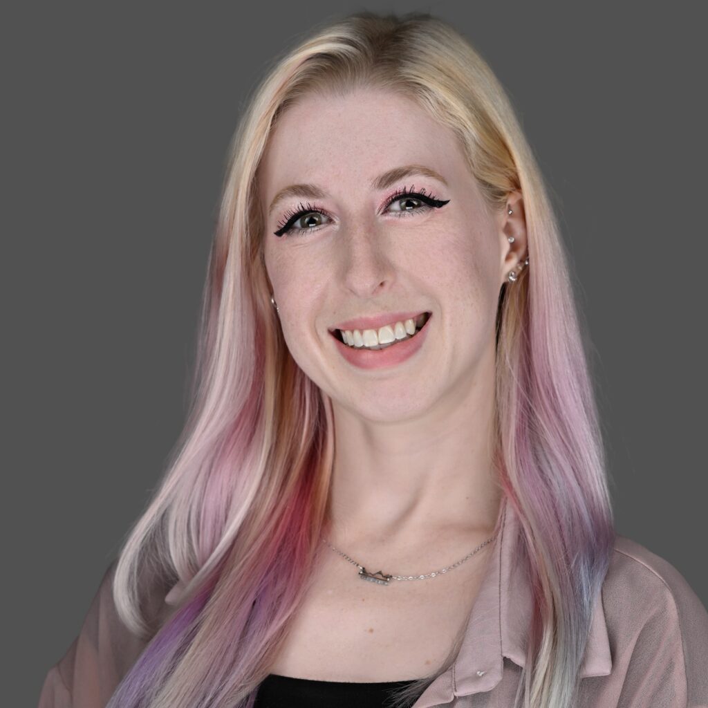 A young woman with long, pastel-colored hair smiles at the camera. She is wearing a light pink blouse, a black top, and a silver necklace, standing against a plain gray background.