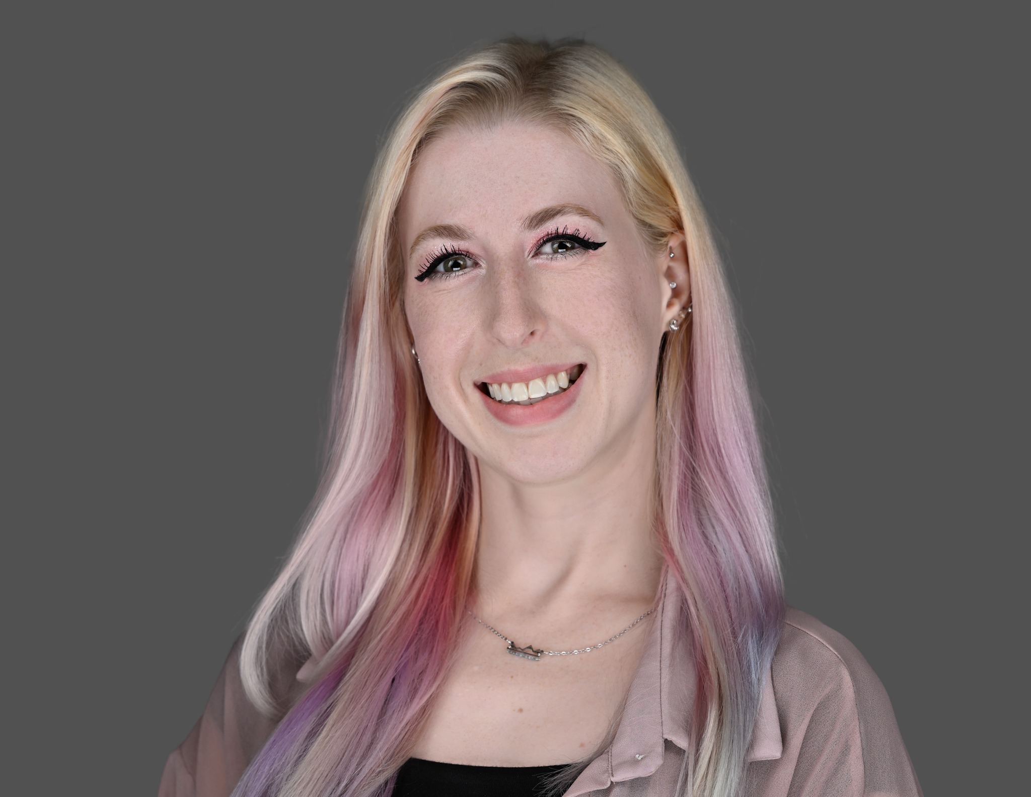 A young woman with long, pastel-colored hair smiles at the camera. She is wearing a light pink blouse, a black top, and a silver necklace, standing against a plain gray background.