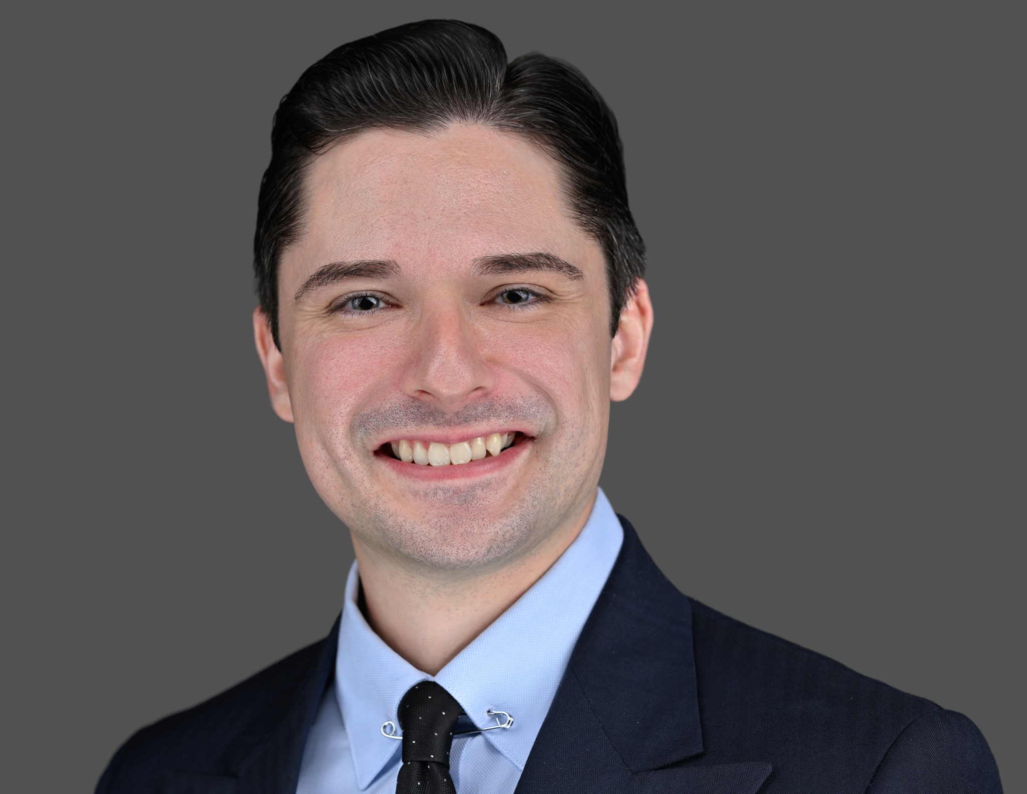 A man with dark hair wearing a navy suit, light blue shirt, and black tie smiles at the camera against a plain gray background.