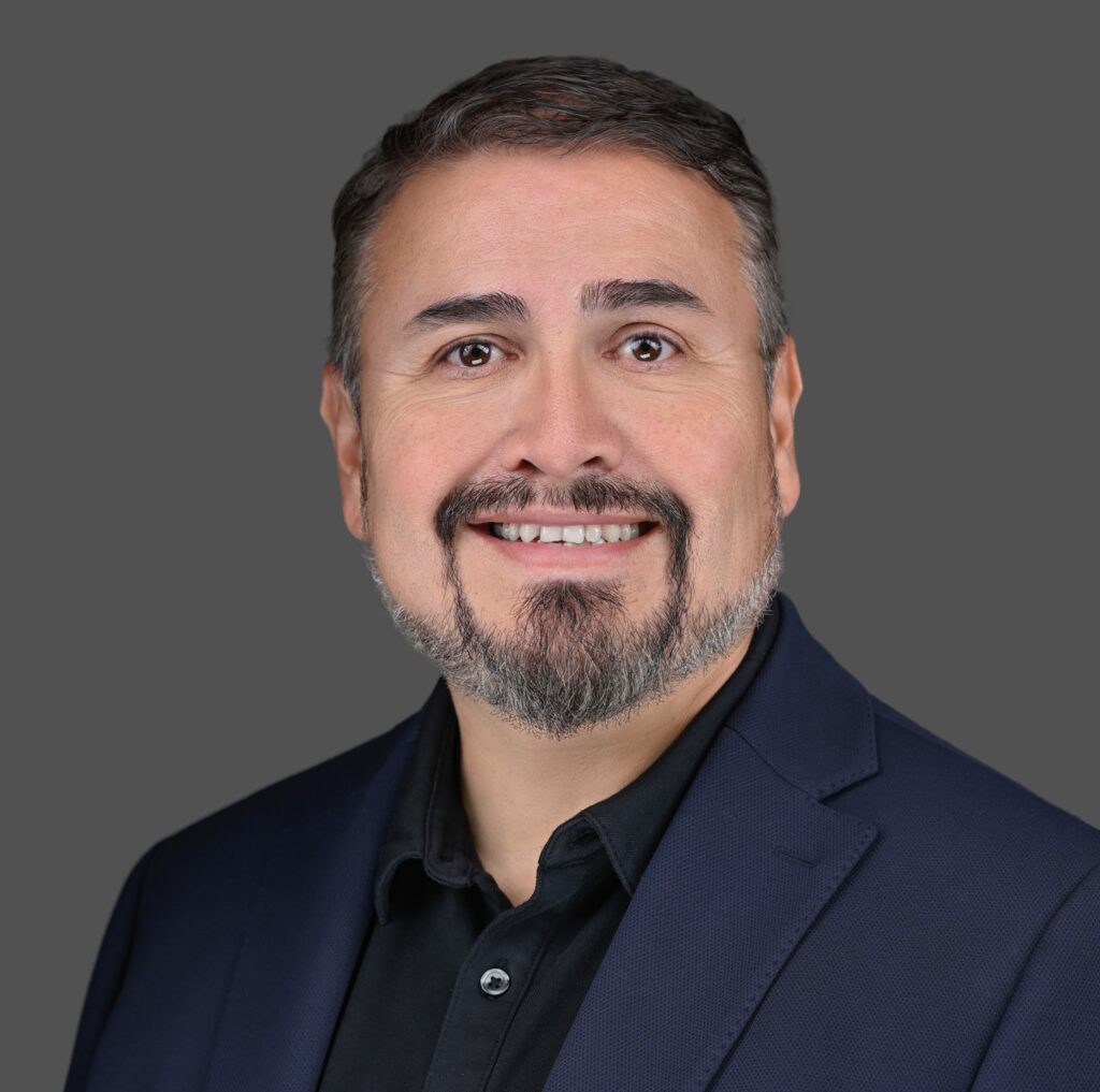 A man with short dark hair, a trimmed beard, and mustache is smiling and wearing a navy blazer and black shirt, posed against a plain gray background.