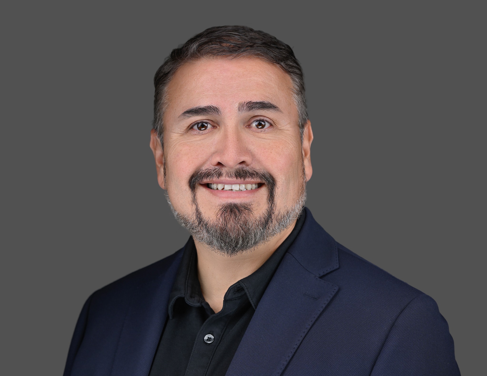 A man with short dark hair, a trimmed beard, and mustache is smiling and wearing a navy blazer and black shirt, posed against a plain gray background.