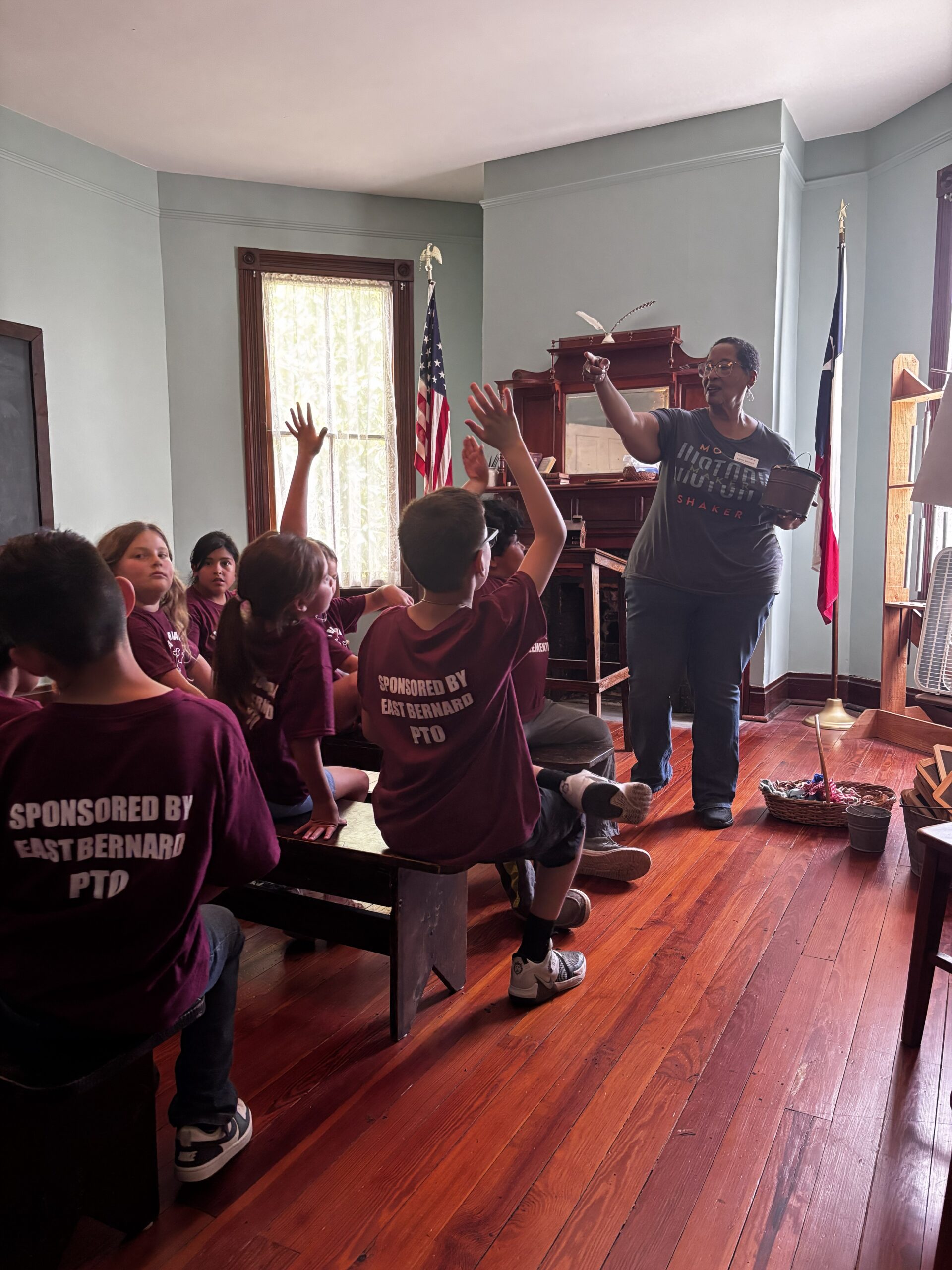 A group of children in matching maroon shirts sit at desks, raising their hands toward a standing adult who is holding a book. An American flag and a Texas flag are beside a bookshelf in the bright classroom.