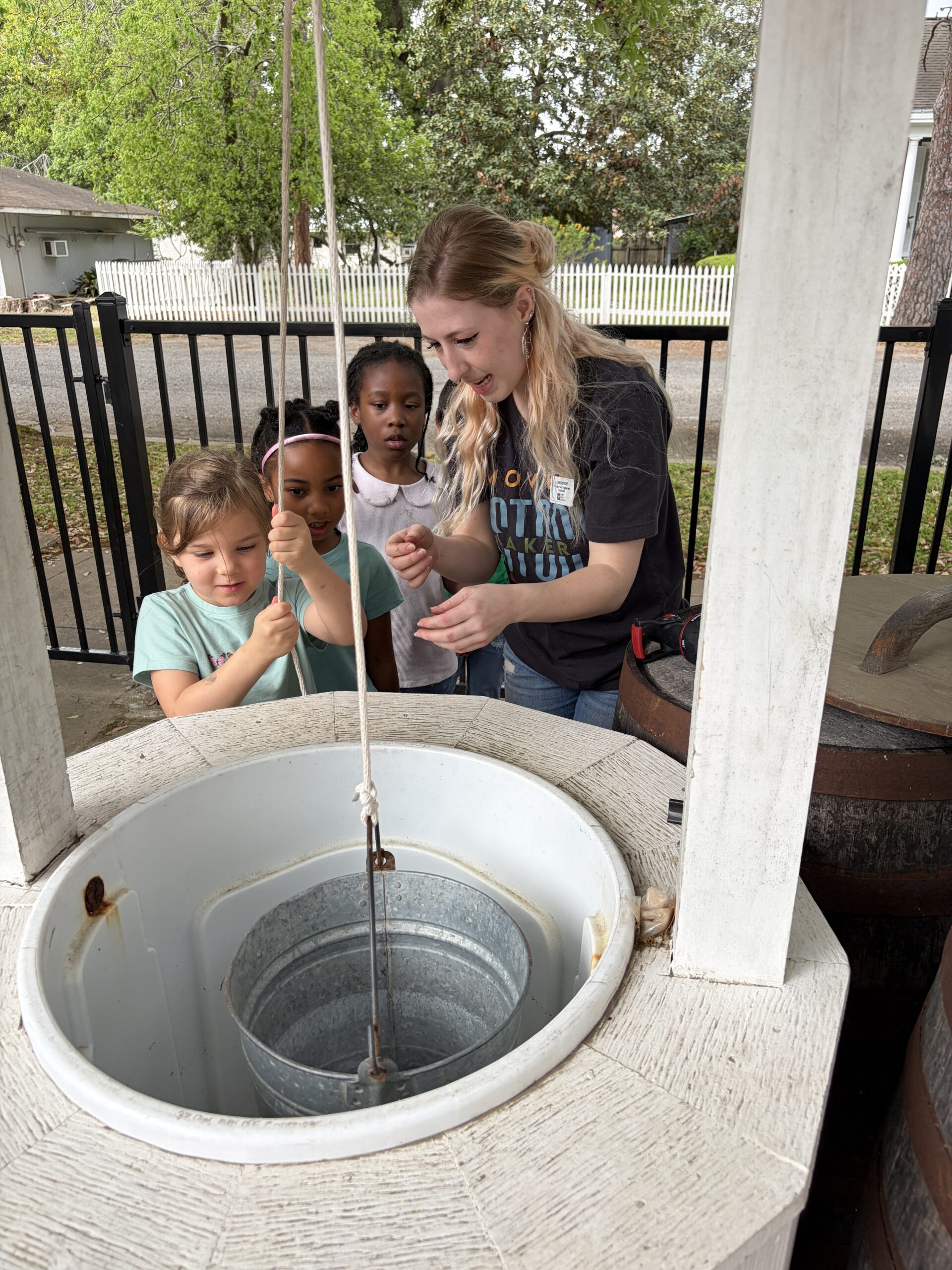 A woman helps three young children draw water from a well using a metal bucket on a rope. They are outdoors near a fence, with trees and a white house in the background.