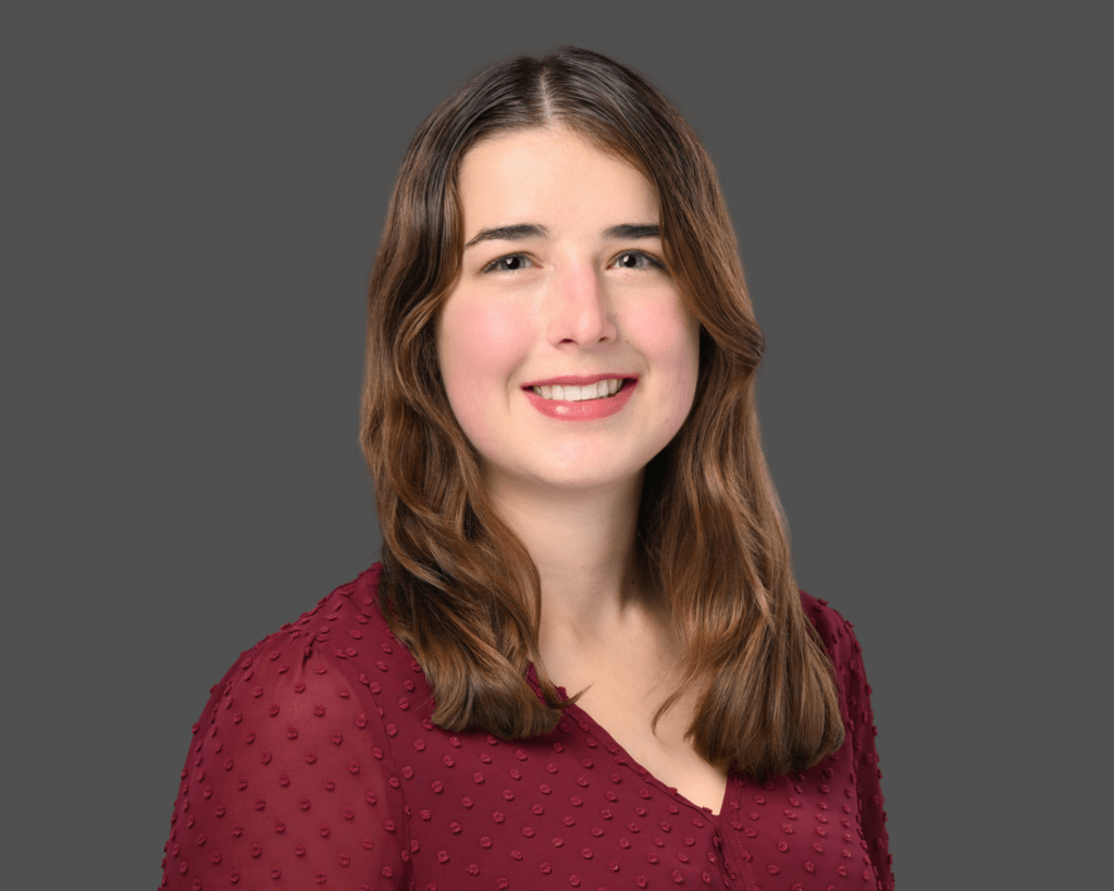 A young woman with long brown hair, wearing a maroon blouse with textured dots, smiles at the camera against a plain gray background.