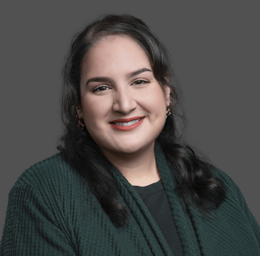 A woman with long dark hair, wearing a dark green cardigan over a black shirt, smiles at the camera against a plain gray background.