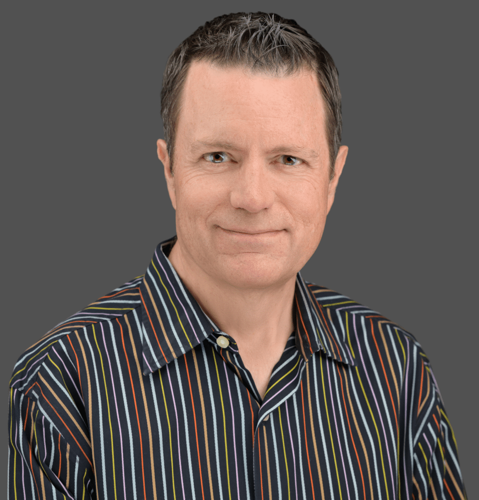 Smiling man with short brown hair, wearing a black shirt with colorful vertical stripes, in front of a plain gray background.