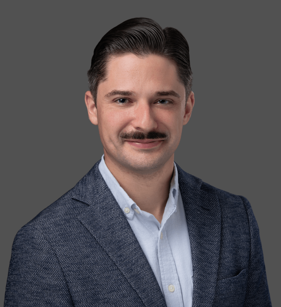 A man with dark hair and a mustache wears a blue textured blazer over a light collared shirt, smiling slightly against a plain gray background.