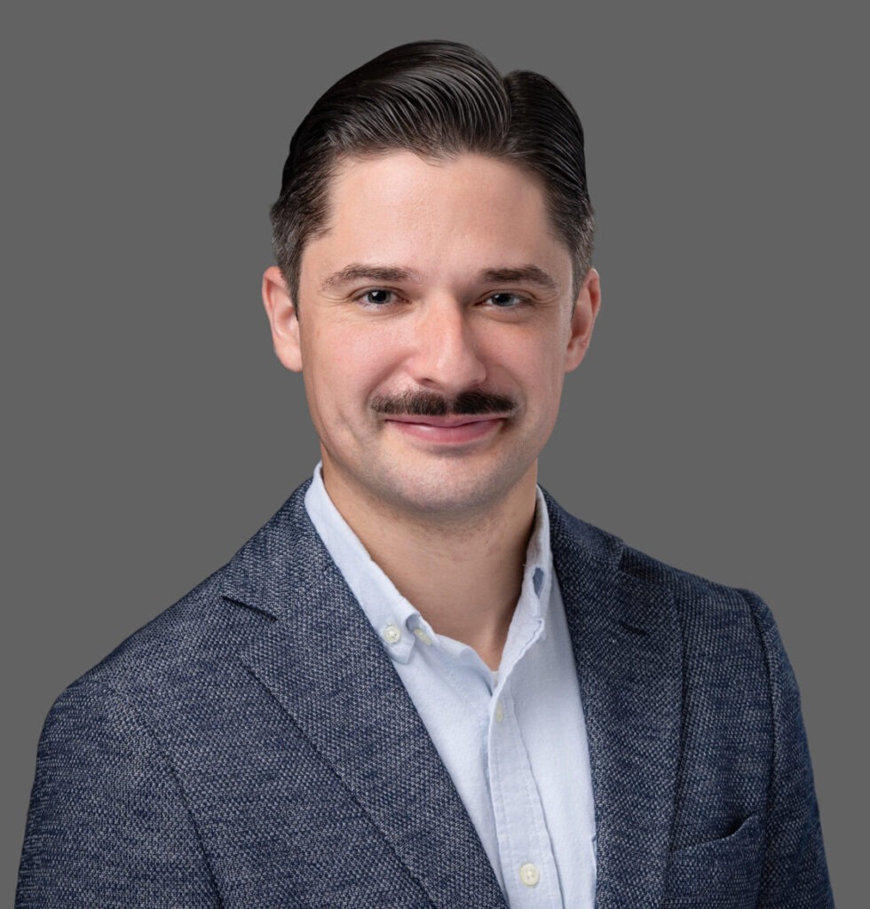 Smiling man with dark hair and mustache wearing a blue blazer and light shirt, posing against a plain gray background.