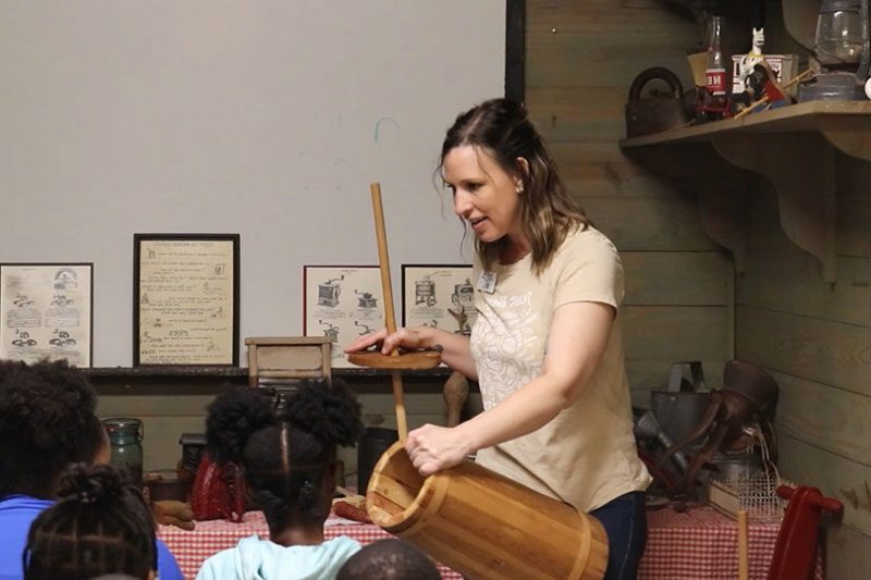 MVI_3244_exported_313 A woman demonstrates how to use a wooden butter churn to a group of children seated at a table in a rustic classroom decorated with vintage items.