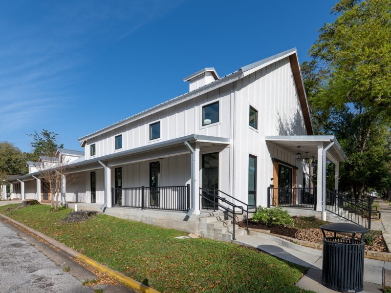 A modern white building with a slanted roof, large windows, and a porch with railings, situated on a corner lot with green grass, trees, and a blue sky above.