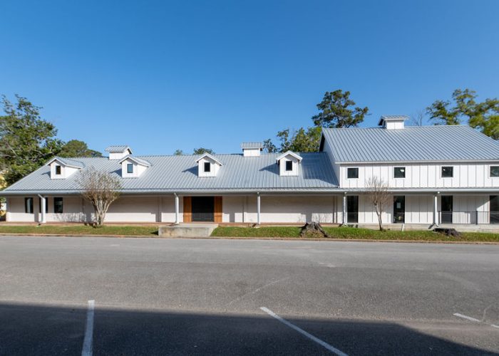 A long, white, single-story building with a metal roof, several dormer windows, and a covered porch, set against a clear blue sky with trees in the background and an empty parking lot in front.