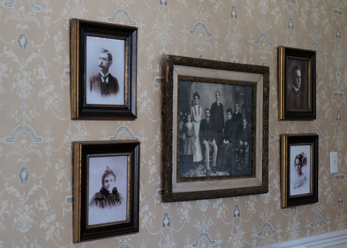 Five framed black-and-white vintage portraits hang on beige patterned wallpaper; four are individual headshots, and the center frame features a group of people.